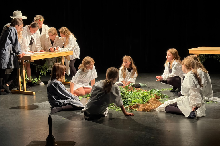 Kinder im Laborkittel und mit Schutzbrillen untersuchen eine große Pflanze auf einer Theaterbühne.